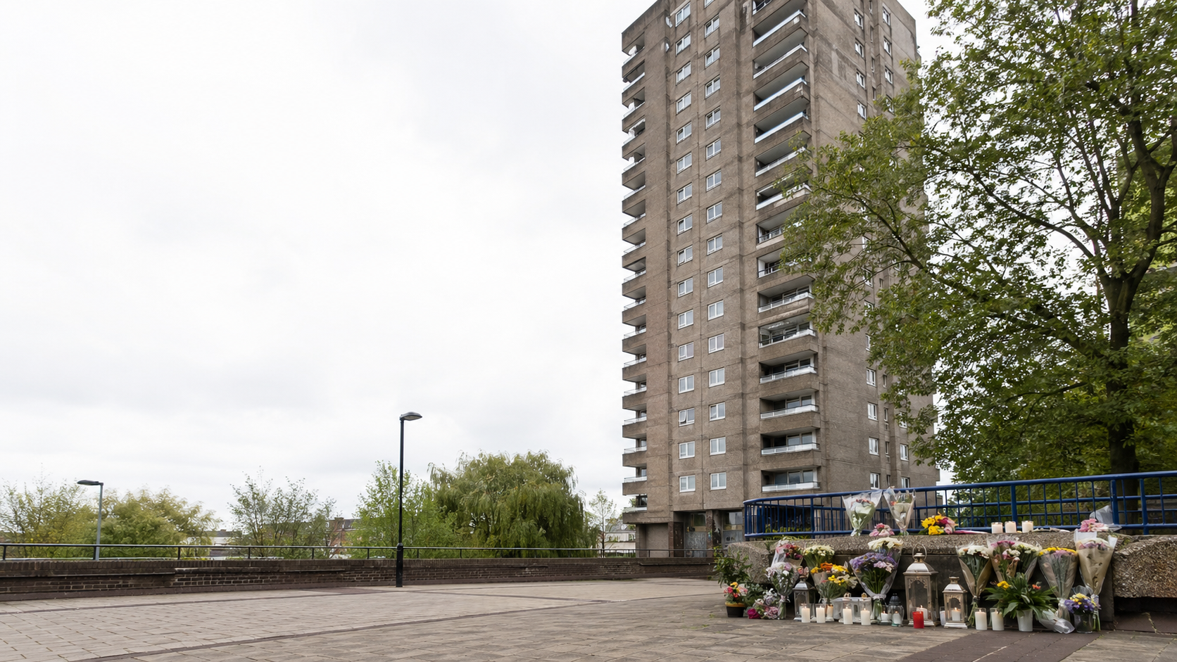 A respectful editorial-style image of a London high-rise residential tower with a small memorial of flowers in the foreground on an overcast day.