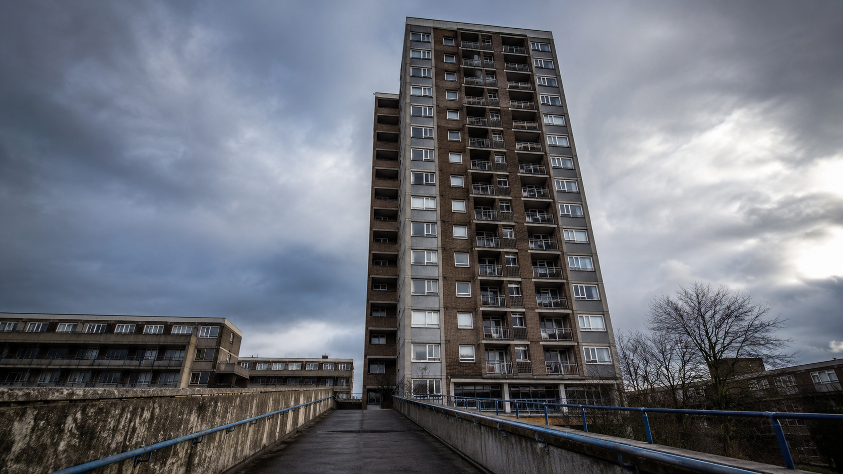 Dramatic low-angle view of a high-rise UK council block with balconies and weathered brickwork under a cloudy sky, representing building safety and fire safety concerns in high-rise housing.