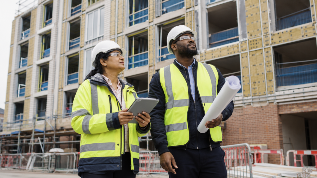 Building control inspectors wearing PPE assessing a high-rise residential construction site with plans and a tablet.